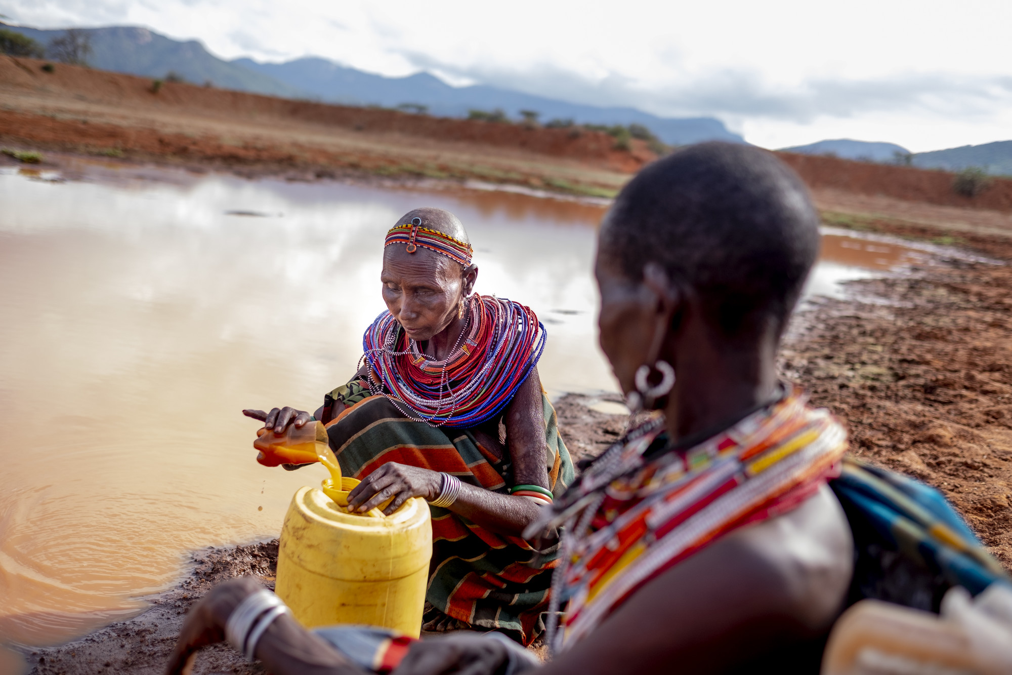 Photo by Christena Dowsett
Nakoyon Lonatu, background, gathers water from a water pan to take home to Lolkuniani. She had measles when she was 4 years old and lost eye-sight in her left eye. It can take her up to two hours each way to collect water and she always has to find someone who is willing to go with her since she can't see well.